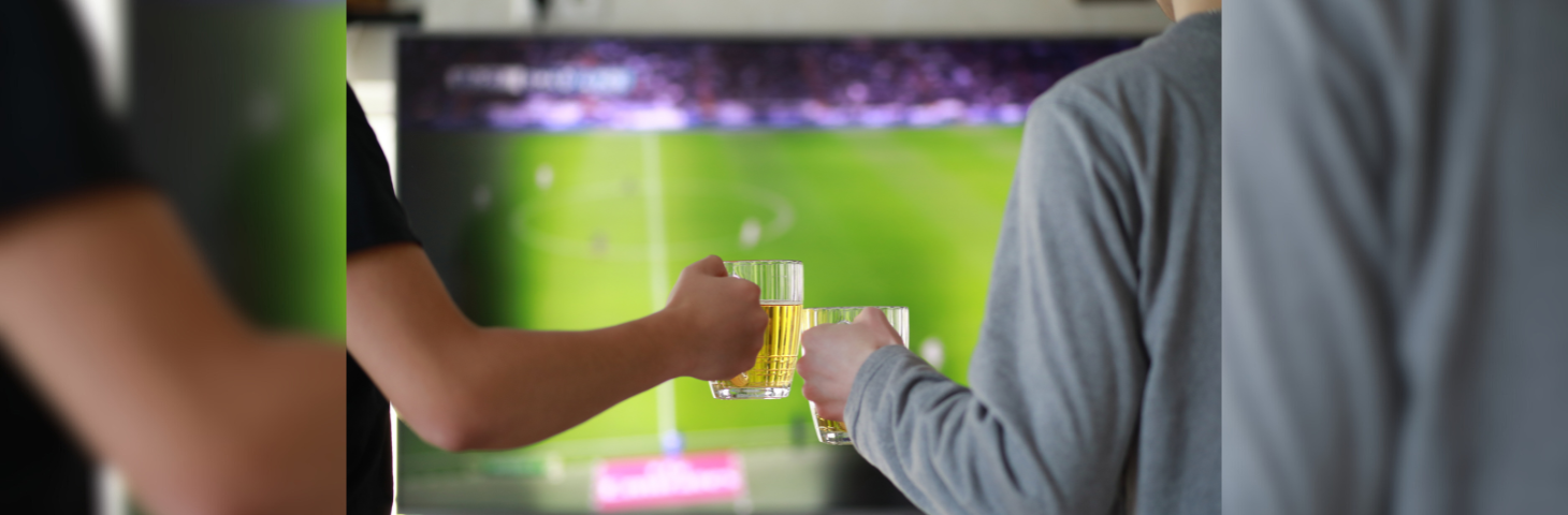Two football fans clinking glasses of beer while watching a game on television.