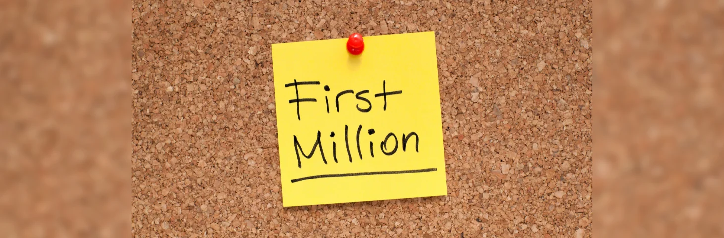 A post it note with 'First Million' written on it in black marker pen pinned to a corkboard.