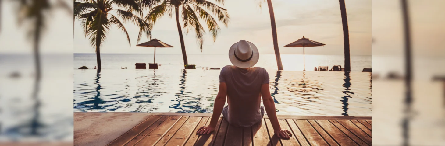 A person on holiday sat on the edge of an infinity pool in a grey-tshirt and cream fedora.