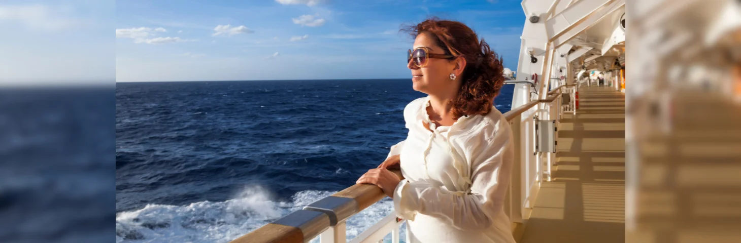 A person in a white shirt and sunglasses stood on the deck of a cruise ship, looking out to sea.