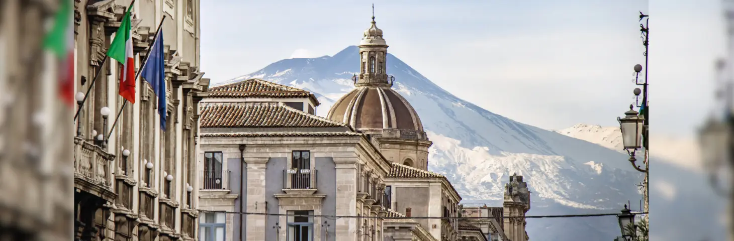 Buildings in Catania with flags adorning the walls in front of a snow-capped mountain