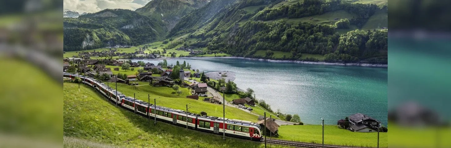 A train traveling along a scenic route next to a lake with mountains in the background. 