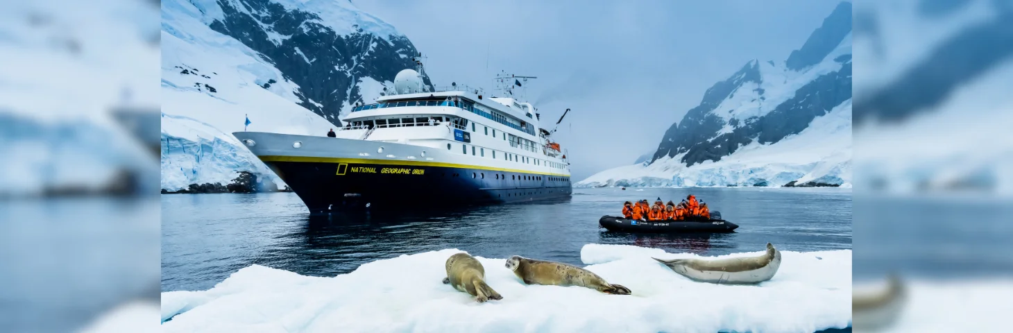 National Geographic Orion expedition ship in Antarctica, with passengers on a smaller Zodiac craft nearby and seals on an ice floe. 