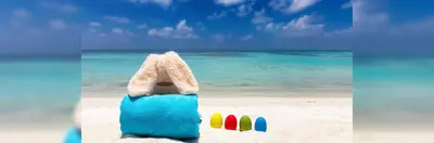 A plush rabbit on a towel on the beach, next to four multi-coloured eggs.