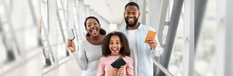A family of three holding their passports while smiling at an airport departure tunnel.