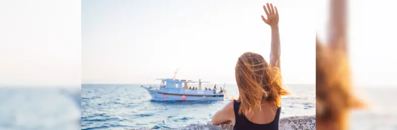 A person with shoulder-length blond hair waving as a boat sails into the distance.