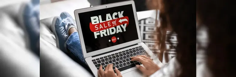 A person with dark hair sat on the couch. Their laptop is open on a screen that says 'Black Friday sale'