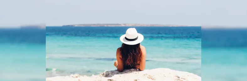 A brown-haired woman in a white hat sunbathing on a rocky shore.