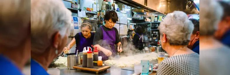 Three chefs in purple aprons preparing Okonomiyaki in Hiroshima, watched by three tourists.