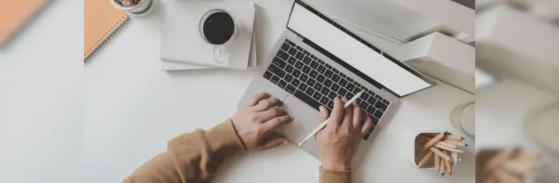Someone in a brown jumper working on an Apple Macbook on a white desk.