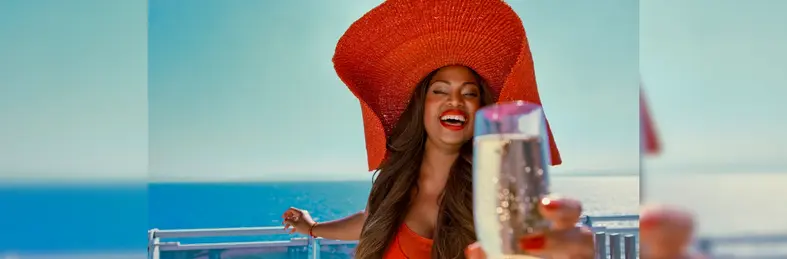 A woman with long brown hair in a red outfit and and red painted nails raising a glass to the camera on a cruise ship.