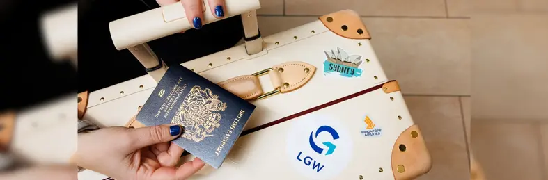 A person with blue painted nails holding a British passport above a suitcase with London Gatwick, Singapore Airlines and Sydney Opera House stickers on.