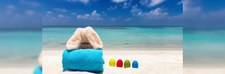 A plush rabbit on a towel on the beach, next to four multi-coloured eggs.