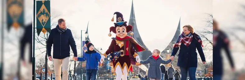 A family of four (two adults and two children) holding hands with Pardoes, Efteling's mascot, outside the park entrance.
