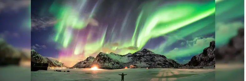 A person stood in front of the Northern Lights over a mountain on Skagsanden Beach in Norway's Lofoten Islands.