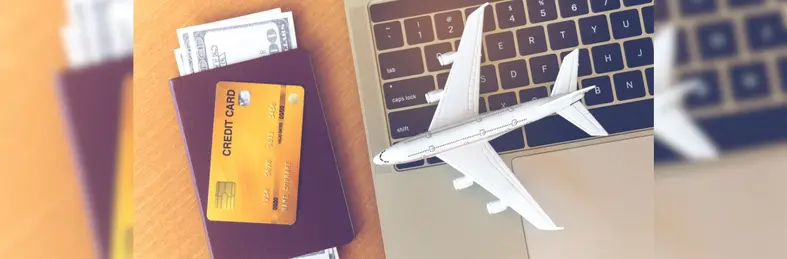An Apple Macbook with a model plane resting on its keyboard next to a passport with American dollars on the inside and a credit card on top.