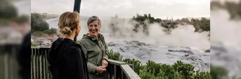 Two women talking to eachother smiling against the backdrop of steam-filled waters in New Zealand