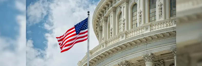 A flag of the USA waving in front of the Capital Building in Washington, D.C., against a cloudy blue sky.