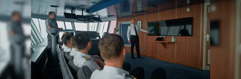 The navigational bridge area of a Holland America Line ship, with crew members undergoing a safety briefing from the Captain. 