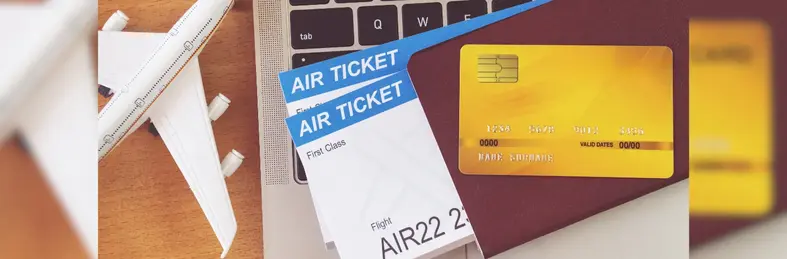 A laptop, miniature airplane, passport containing two First Class flight tickets, and golden rewards card placed on a wooden table.