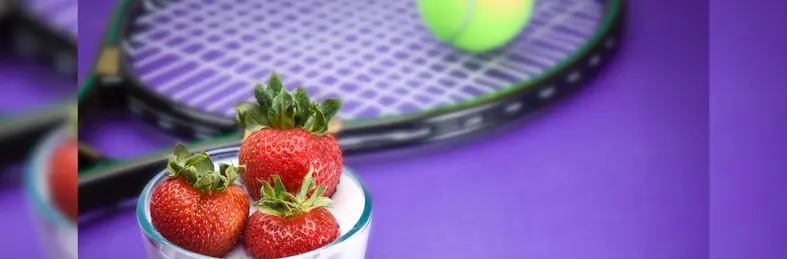 A glass dish of strawberries & cream in front of a tennis racquet and ball.