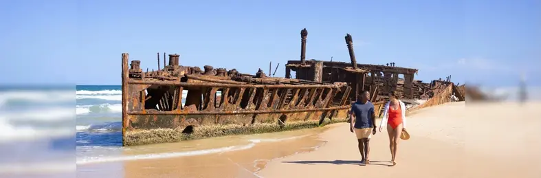 Two people, one in a blue t-shirt and beige shorts and one in a red swimsuit, walking past the wreck of the World War I hospital ship SS Maheno on Fraser Island/K'gari in Queensland, Australia.