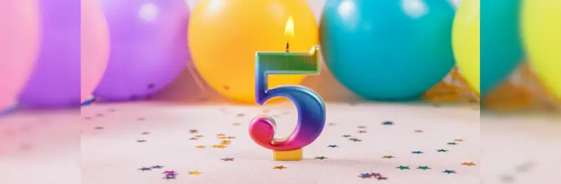 A rainbow-coloured 5 candle in front of balloons with confetti scattered on the table.