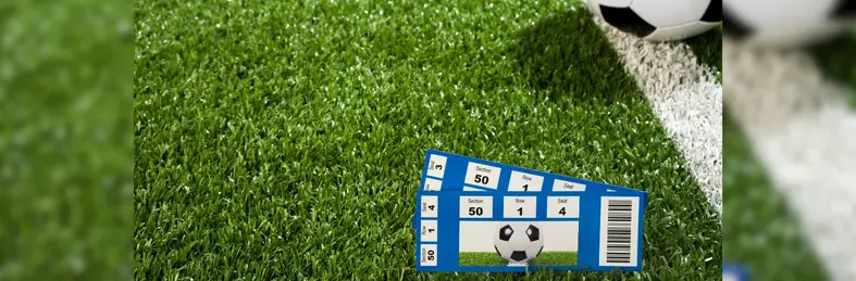 Two blue tickets to a football game on an astro turf pitch, with a football resting next to them.
