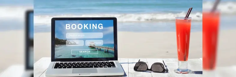 An Apple Macbook, sunglasses and cocktail on a white bench on the beach. The laptop is open on a 'BOOKING' page to manage a holiday booking.