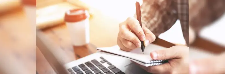 man making notes with a laptop in front of him and a coffee on a desk