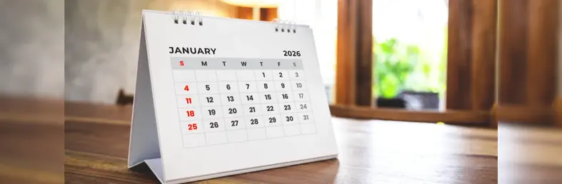 A 2026 calendar, open on the January page, on top of a wooden desk.